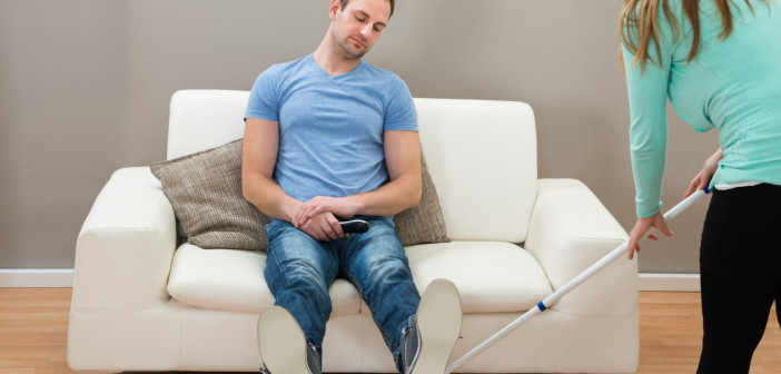 Woman Cleaning Floor With Mop While Man Sitting On Sofa